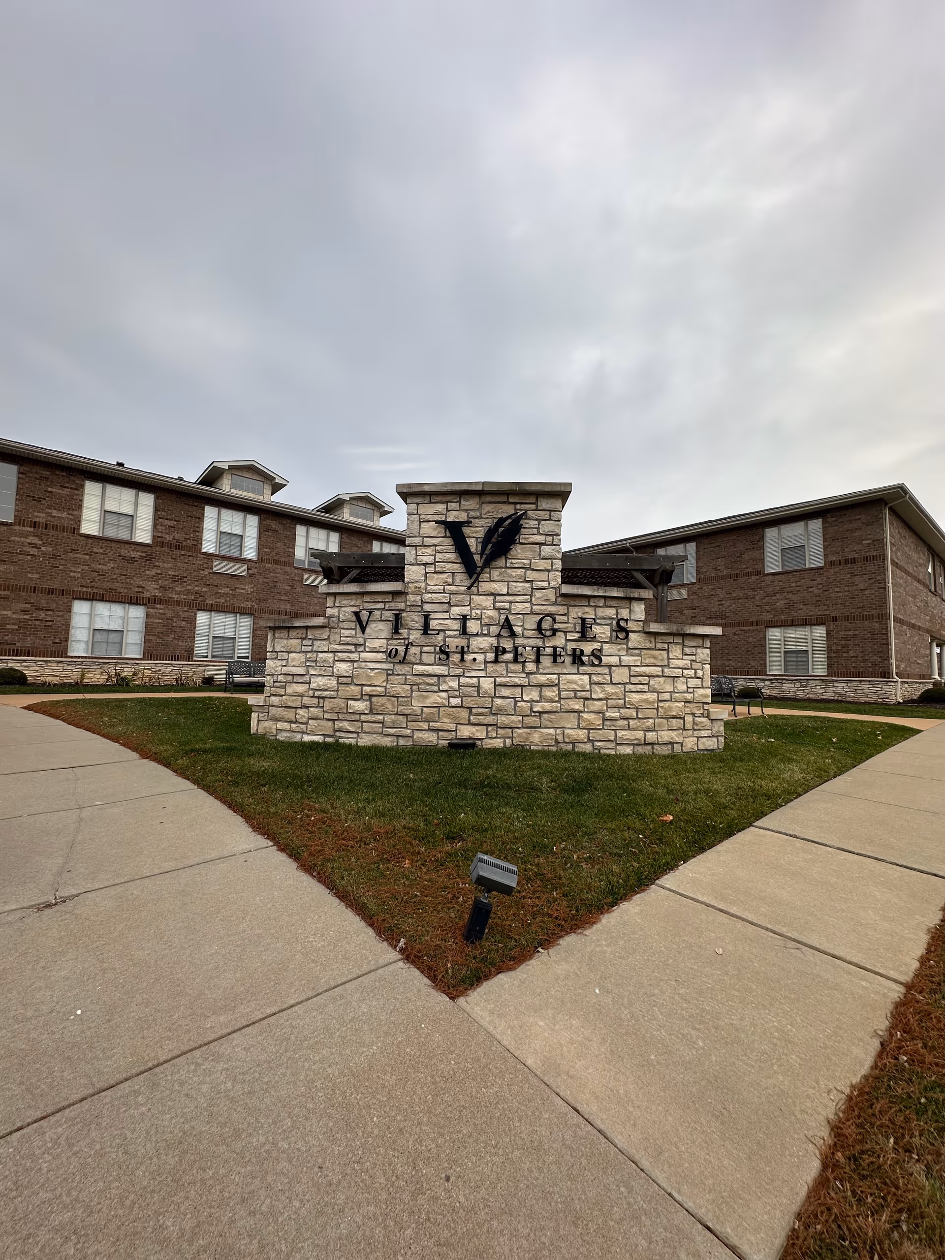 Stone monument sign that reads "Villages of St. Peters" in front of a two-story brick building under a cloudy sky.