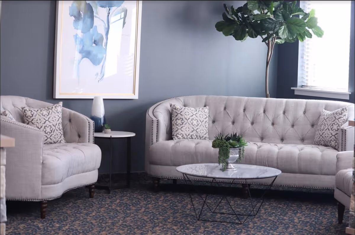 A cozy living room area with a tufted beige sofa and matching armchairs, each adorned with patterned throw pillows. A round marble coffee table with a small green plant centerpiece sits in front of the sofa. A side table with a decorative vase is placed between the sofa and armchair. A large leafy plant stands near a window with blinds, and a framed abstract blue and white painting hangs on the gray wall behind the sofa.