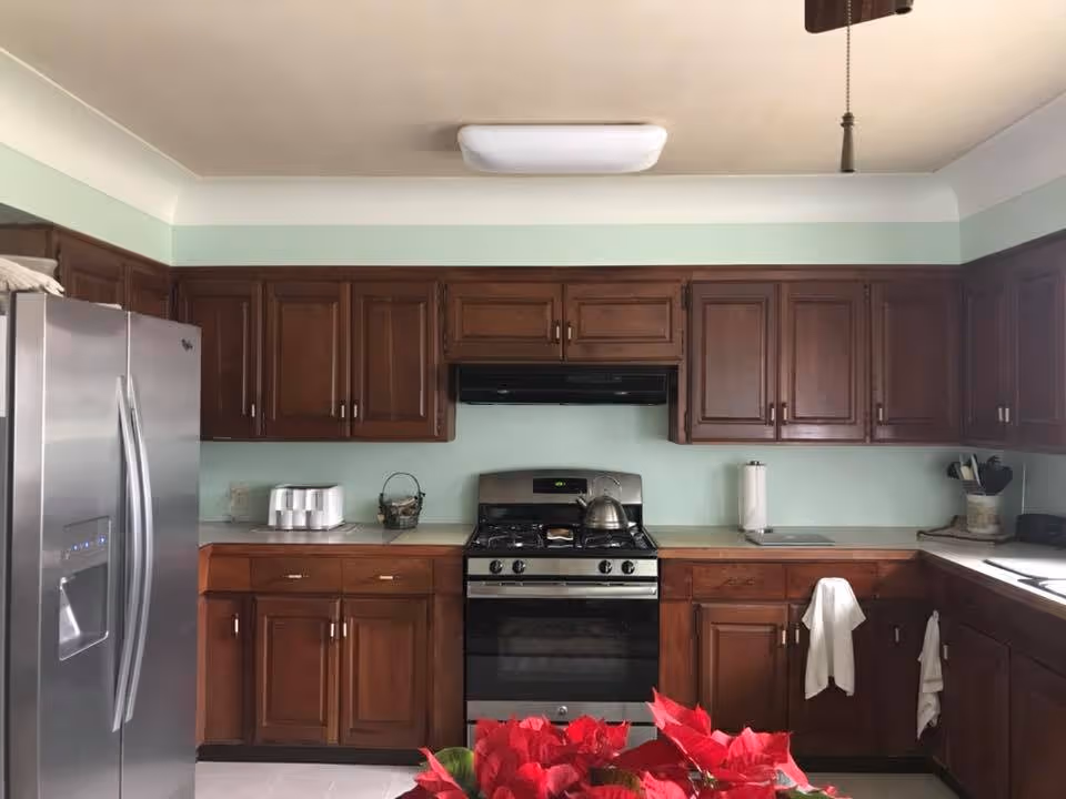 A kitchen with dark wooden cabinets, a stainless steel refrigerator on the left, a stainless steel gas stove with a kettle on it in the center, and a countertop with various kitchen items including a paper towel holder and utensils. The walls are painted light green and there is a ceiling light fixture.