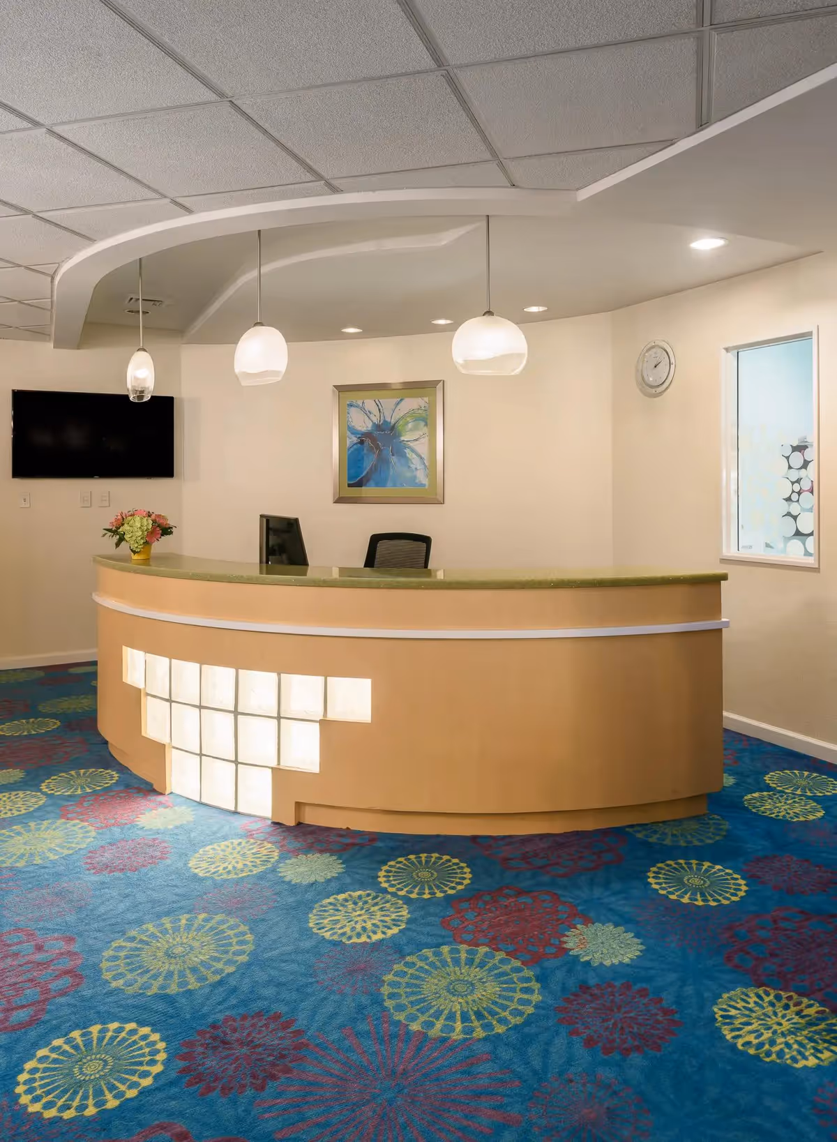 Reception area with a curved wooden desk featuring illuminated square panels, a computer monitor, a black office chair, and a small flower arrangement. The room has a colorful carpet with circular patterns in blue, yellow, and red. Three pendant lights hang from the ceiling above the desk. A wall-mounted TV, a framed abstract painting, a clock, and a window with frosted glass are visible on the walls.