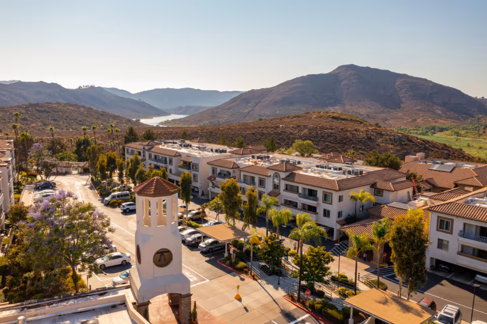 Aerial view of Casa de las Campanas senior living facility with multiple buildings featuring terracotta roofs, surrounded by trees and parked cars. In the foreground, there is a white bell tower structure. The background shows dry hills and a river winding through the valley under a clear sky.