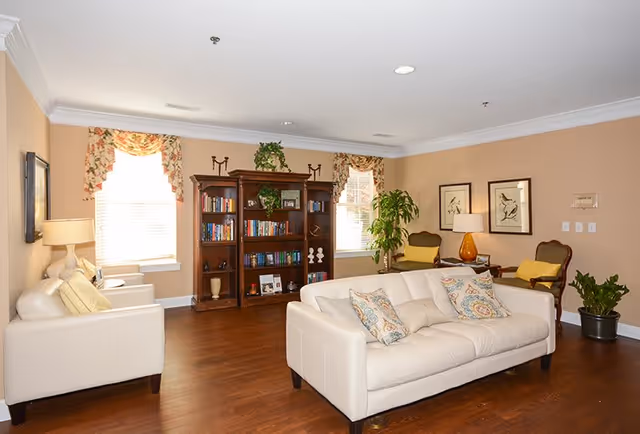 A cozy living room with two white sofas adorned with patterned cushions, two armchairs with yellow pillows, a wooden bookshelf filled with books and decorative items, two windows with floral curtains, framed artwork on the wall, a table lamp with an orange base, and several potted plants on a wooden floor.