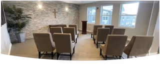 A small chapel or meditation room with two rows of beige upholstered chairs facing a wooden podium. The back wall is decorated with light-colored stone and a cross mounted above. Large windows on the right side let in natural light, and there is a tall green plant in the left corner.