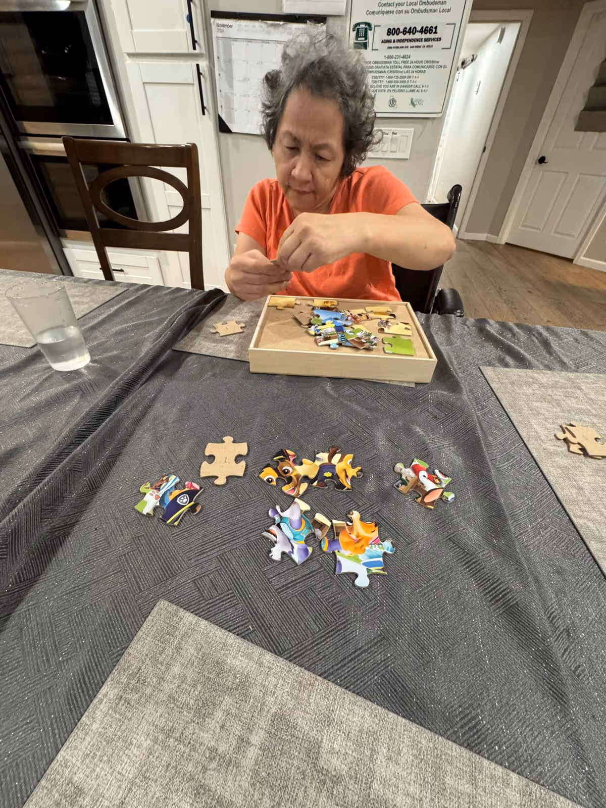 An elderly woman with short curly hair wearing an orange shirt is sitting at a table working on a colorful jigsaw puzzle. The table has a dark textured tablecloth and a glass of water. The background shows a kitchen area with white cabinets and a wall calendar.