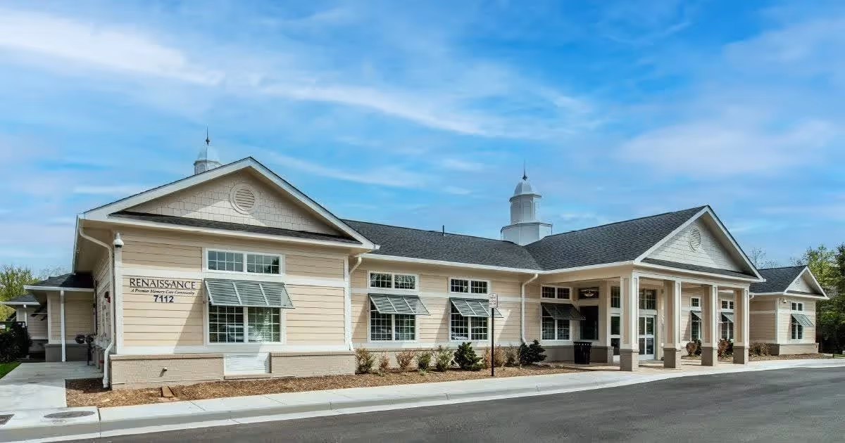 Exterior view of Renaissance of Annandale senior living facility building with beige siding, multiple windows with awnings, and a covered entrance under a blue sky with some clouds.