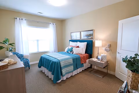 Furnished bedroom with a teal-accented bed, nightstand, dresser, and a window with white curtains.