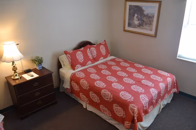 A bedroom with a bed covered in a red and white patterned bedspread and matching pillows. Next to the bed is a wooden nightstand with a brass lamp, a small potted plant, and a remote control. A framed painting hangs on the wall above the bed, and there is a window with blinds letting in natural light.