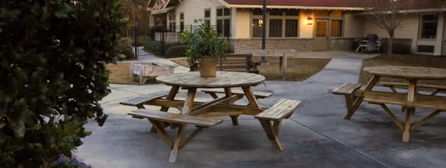 Outdoor patio area with round wooden picnic tables and benches on a concrete surface. There is a potted plant on one of the tables. In the background, there is a building with windows and exterior lights turned on, surrounded by a fenced garden area and some trees.