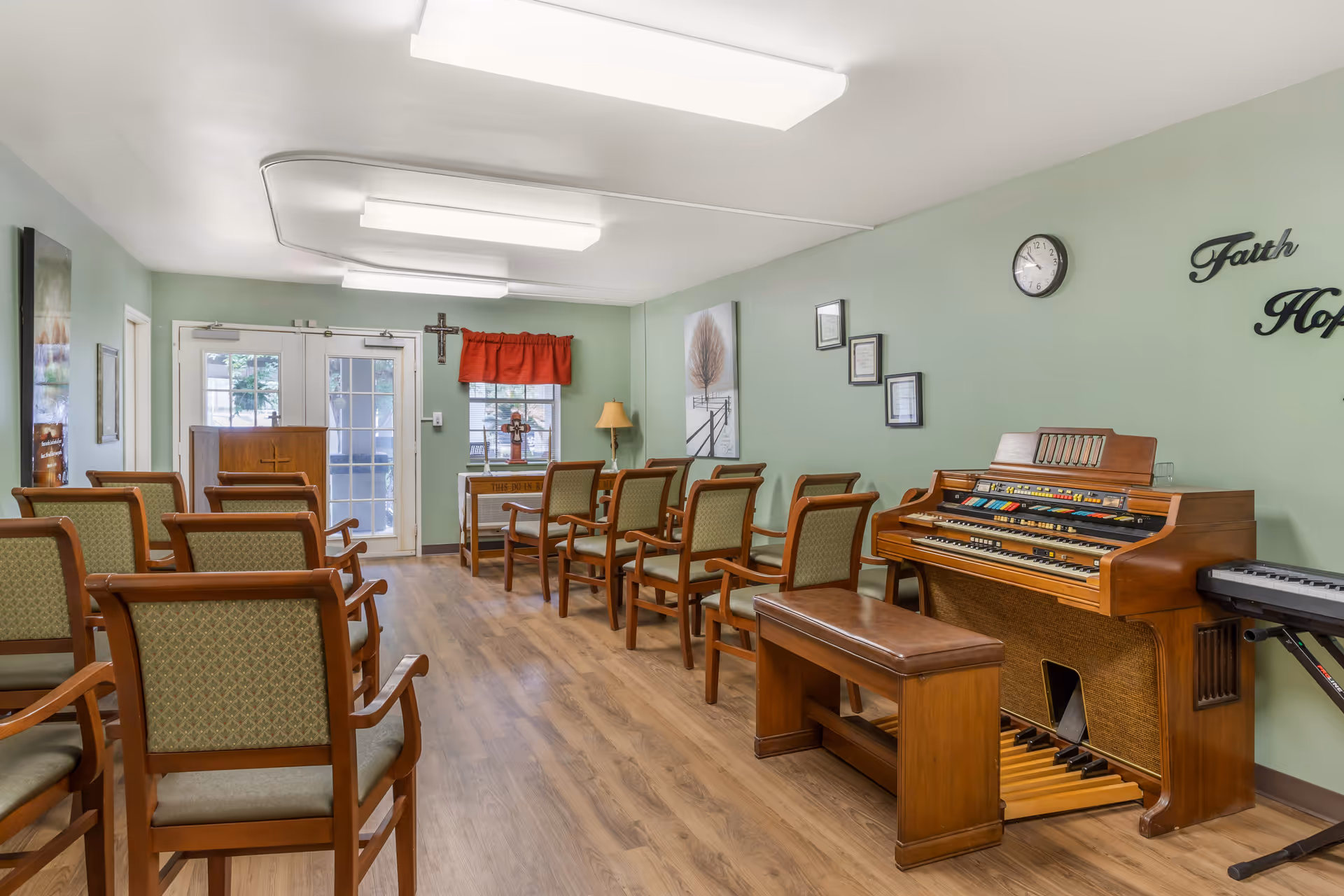 A small chapel or prayer room with wooden chairs arranged in rows facing a wooden podium and a small altar with crosses. The room has light green walls, a wooden floor, a clock on the wall, and a vintage organ with a bench on the right side. There are framed pictures and inspirational words on the walls, and double glass doors at the back letting in natural light.