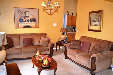 A cozy living room with two brown upholstered sofas adorned with decorative pillows, a wooden coffee table with decorative items, a side table with a lamp, and framed artwork on yellow walls.