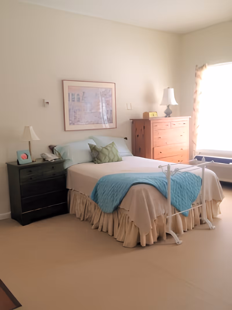 Bright bedroom featuring a made bed with a blue throw and bed rail, two nightstands, a wooden dresser, and a window letting in sunlight.