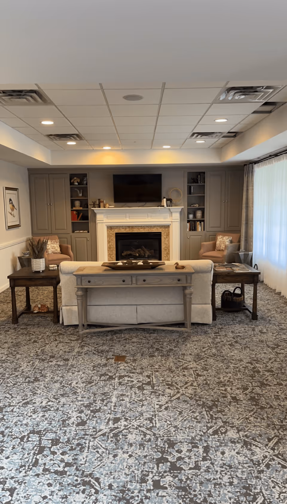 A cozy living room with a patterned carpet, a beige sofa facing a white fireplace with a mounted TV above it. On either side of the fireplace are built-in shelves and cabinets. Two armchairs with cushions are placed near the fireplace, and side tables with decorative items flank the sofa. Large windows with sheer curtains allow natural light into the room.