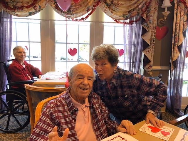 Three elderly people at a decorated dining table—two posing together in the foreground while another sits in a wheelchair near a window.