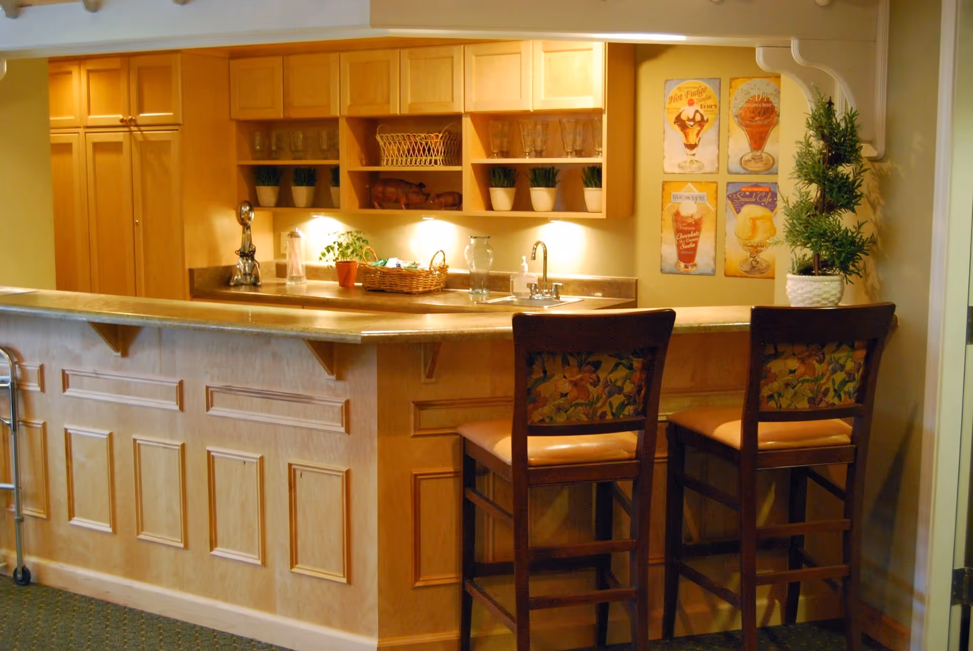 A cozy kitchen area with a wooden counter and two high chairs with floral upholstery. The counter has a sink, a basket, a plant, and some glassware on shelves above. Four vintage-style posters of ice cream desserts are displayed on the wall next to a potted plant.