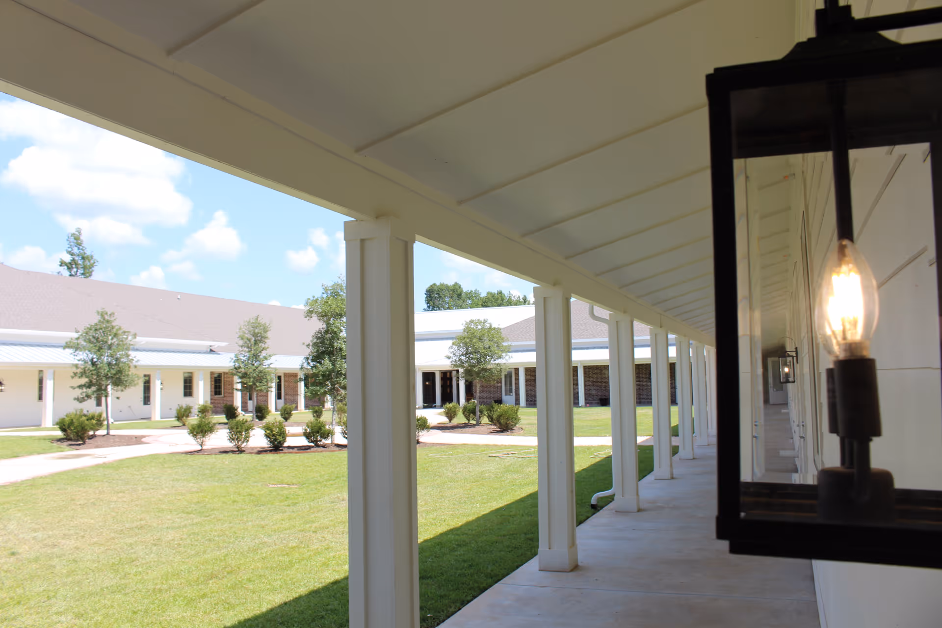 Covered walkway with white columns and a hanging lantern light fixture, leading to a building with a light-colored exterior and a brown roof, surrounded by green grass and small trees under a blue sky with some clouds.