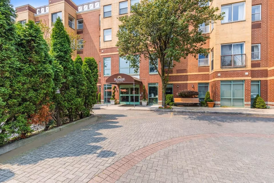 The front entrance of a Sunrise senior living building with a brick facade, awning, trees and a paved circular driveway.