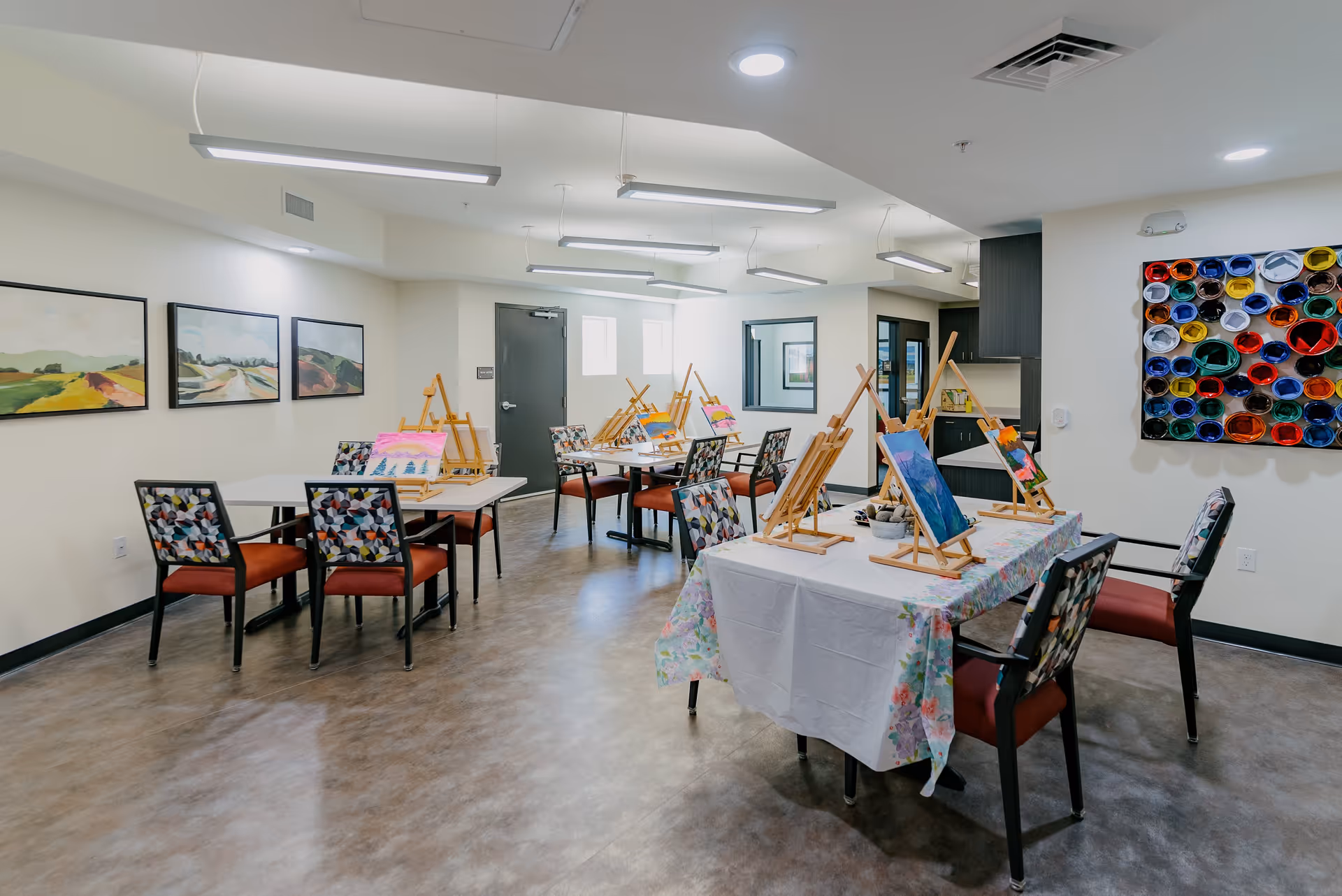A bright and spacious art activity room with multiple tables and chairs arranged for painting. Each table has easels with colorful paintings in progress. The walls are decorated with landscape paintings and a colorful circular art piece made of rolled paper tubes. The room has modern ceiling lights and a clean, polished floor.