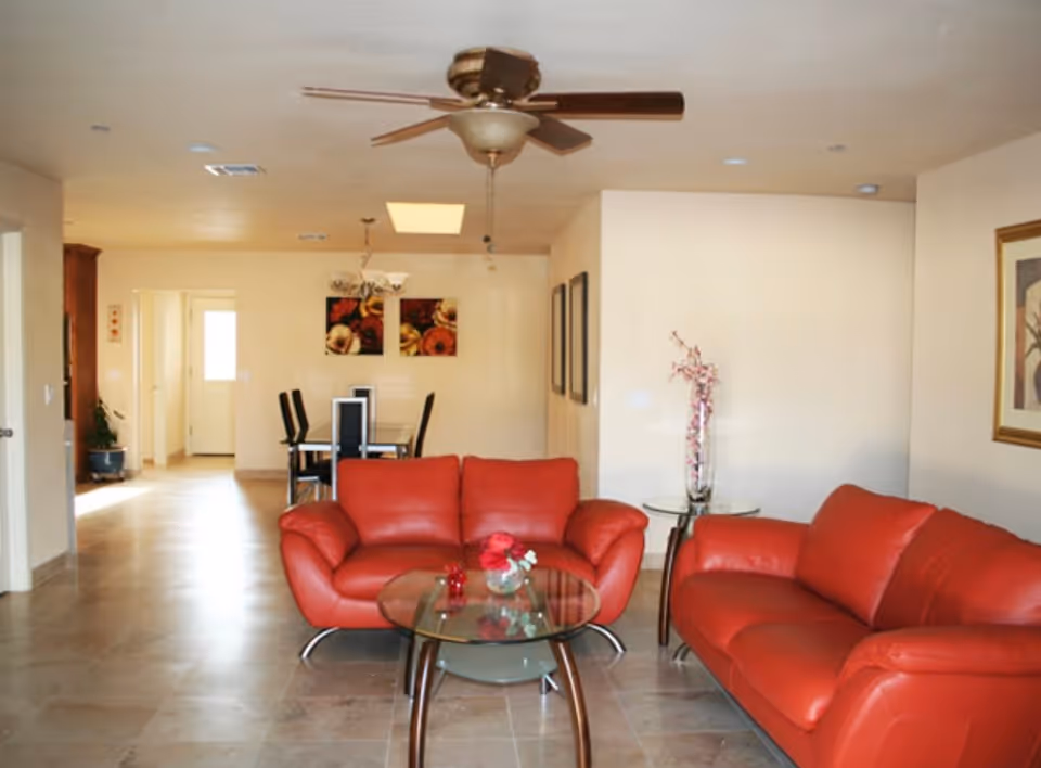 A living room with two red leather sofas facing each other and a glass coffee table in between. Behind the sofas is a dining area with a table and six chairs. The room has beige walls, a ceiling fan with light, and framed artwork on the walls. There is a small side table with a vase of flowers next to one sofa.