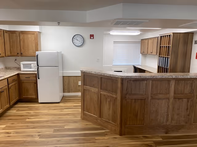 Interior view of a kitchen area with wooden cabinets, a white refrigerator, a microwave, a clock on the wall, and a countertop with a wood-paneled base. The floor is wood-patterned, and there is a window with blinds in the background.