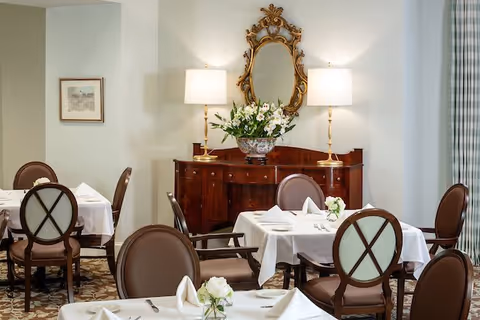 A dining room with several tables covered in white tablecloths, each set with napkins, cutlery, and small flower arrangements. The room features elegant wooden chairs with cushioned seats and backs. Against the wall is a wooden sideboard with two lamps and a decorative mirror above it. A framed picture hangs on the adjacent wall, and a window with patterned curtains is partially visible.