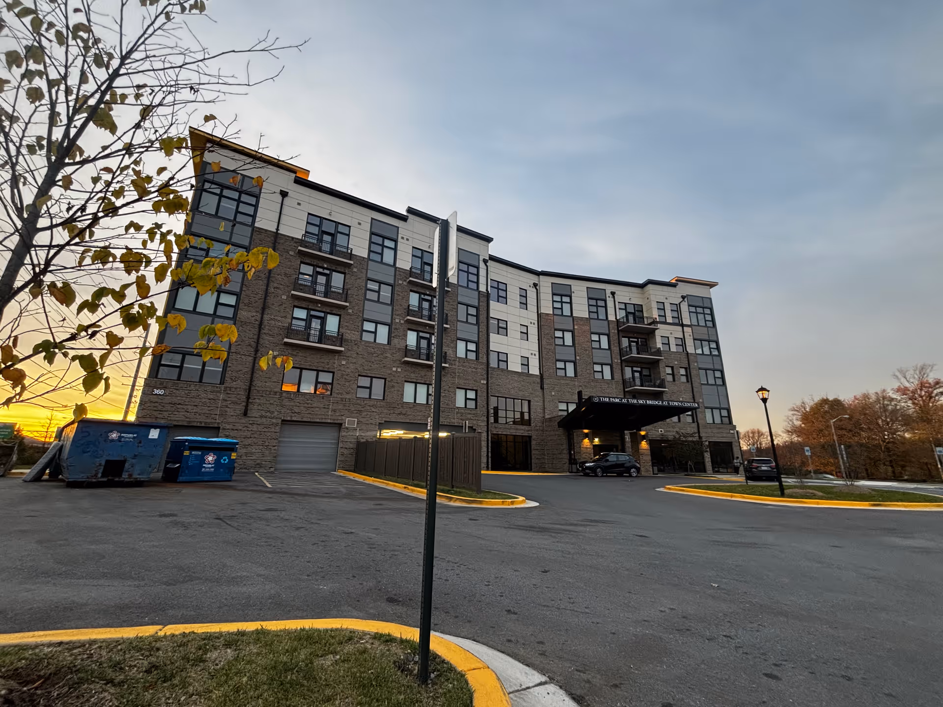 Front exterior of a multi-story senior living building with a covered entrance, parking lot, and a tree at sunset.