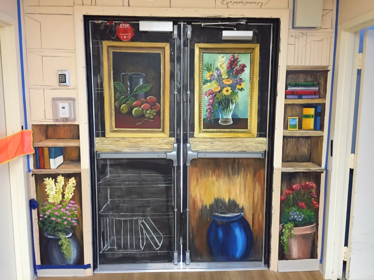 Double doors painted with artistic murals including framed paintings of fruit and flowers, a blue vase, and bookshelves with books and flower pots on either side. The surrounding wall is painted to extend the bookshelf and floral theme.