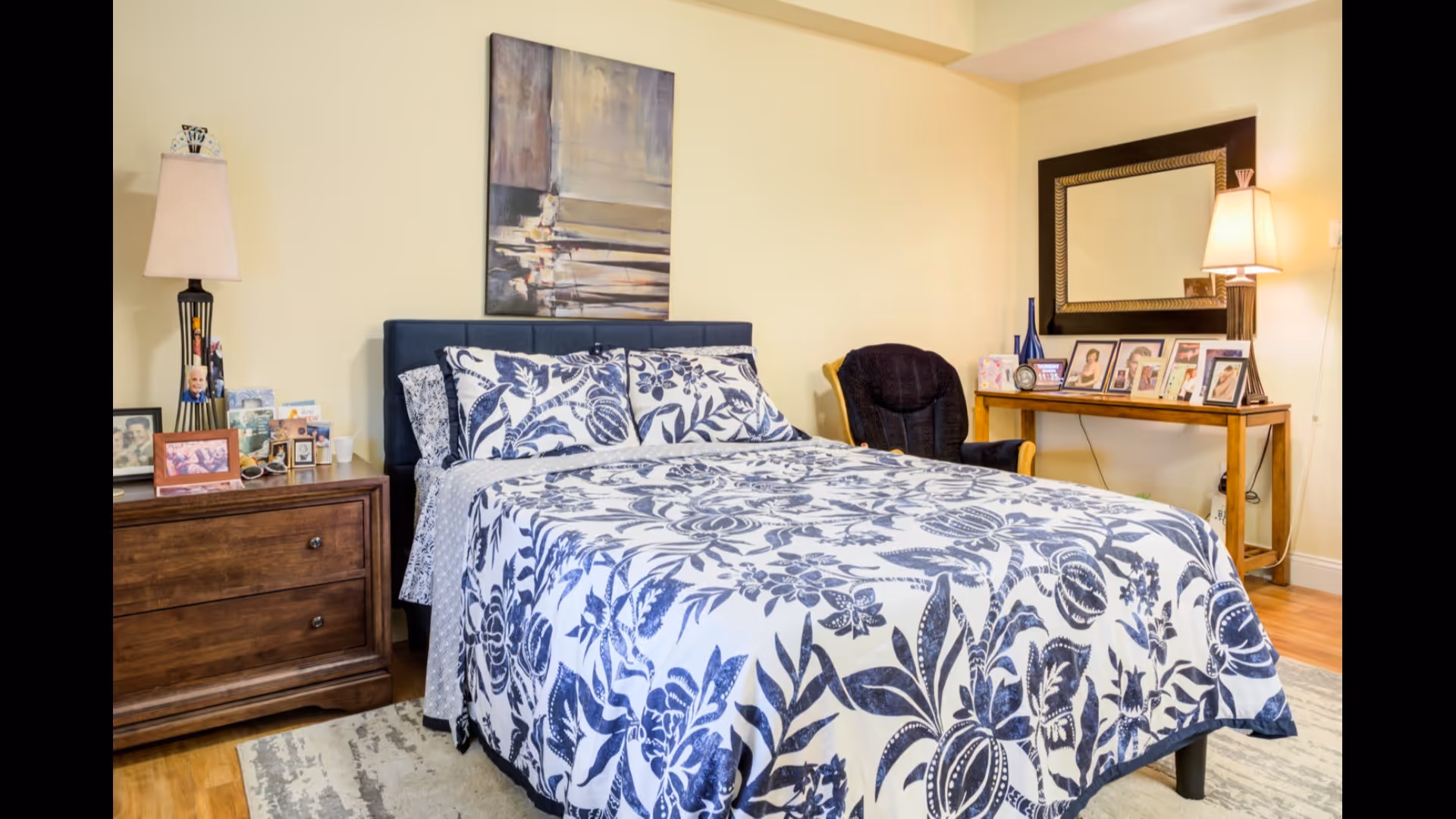 A bright bedroom with a queen bed dressed in blue-and-white floral bedding, wooden nightstands, lamps, framed photos, a mirror, and a chair.