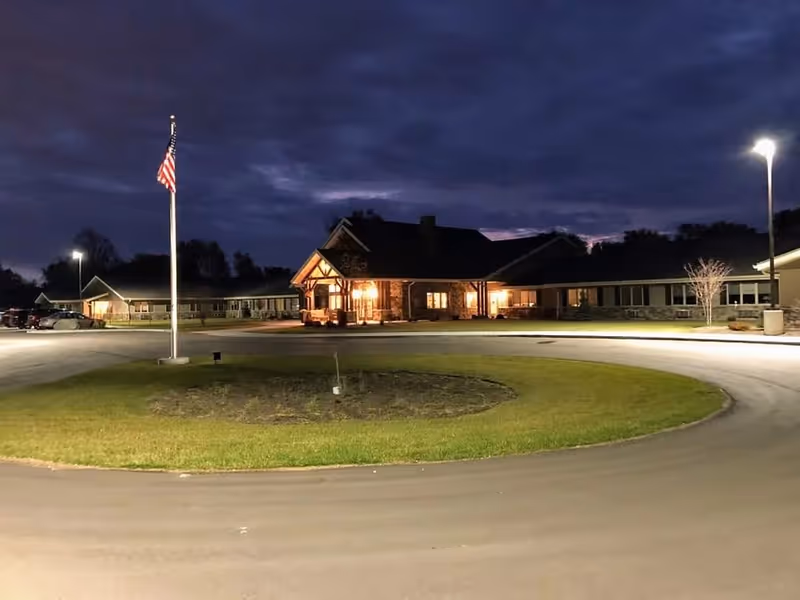 Front entrance of a single-story retirement facility illuminated at dusk with a circular driveway and an American flag in front.