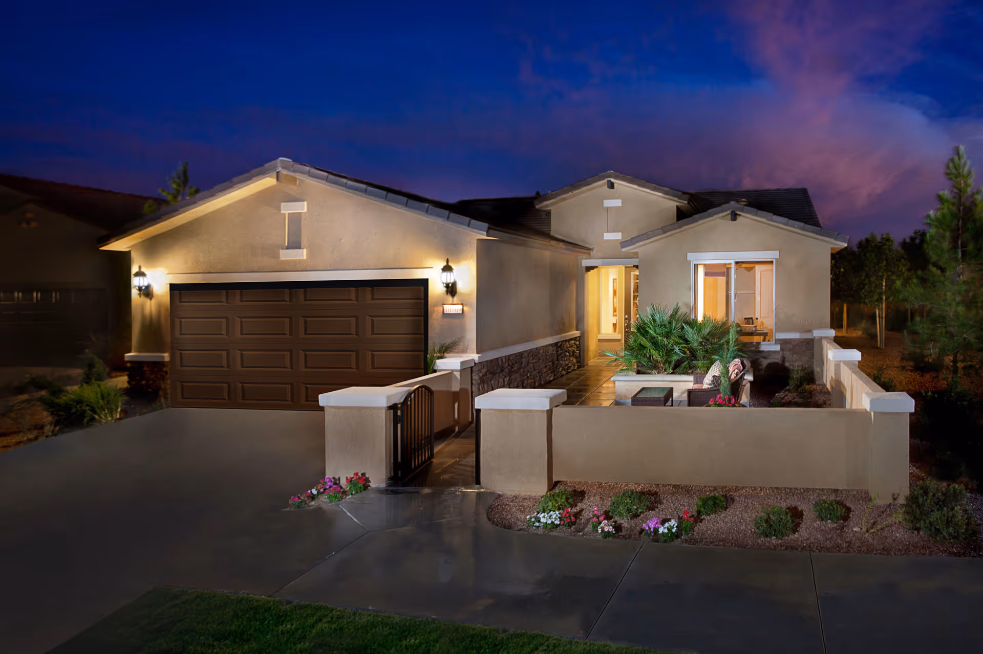 Exterior nighttime view of a single-story house with a two-car garage, illuminated porch lights, a small gated patio with outdoor seating, and landscaped front yard with flowers and shrubs.