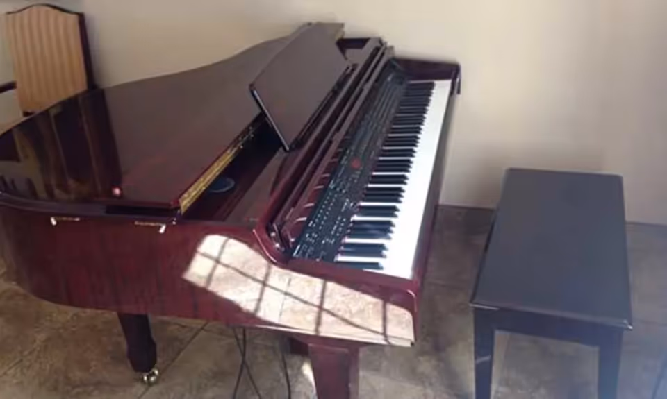 A polished wooden grand piano with its lid partially open, accompanied by a black piano bench, placed on a tiled floor in a room with plain walls.