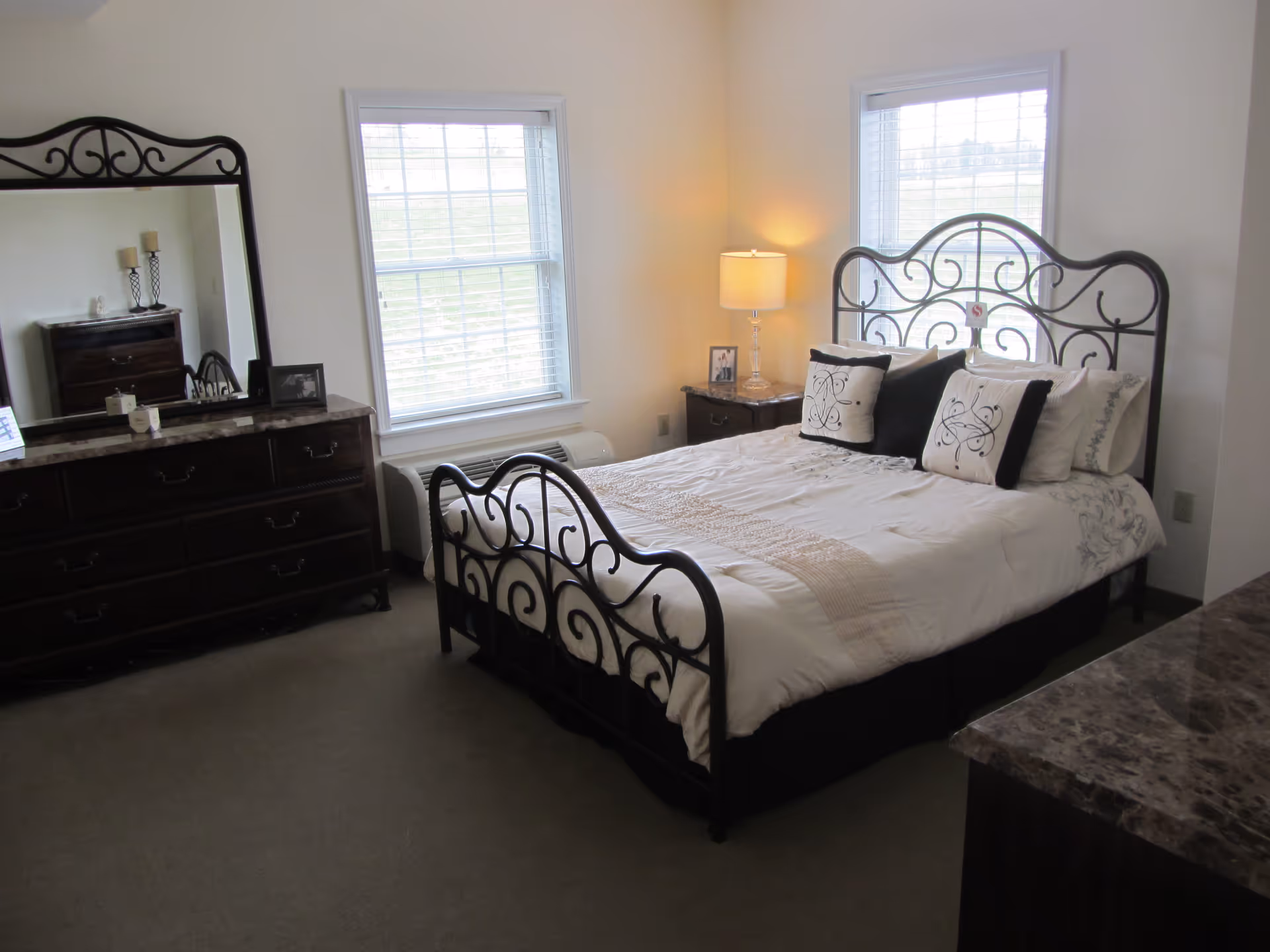 A bedroom with a decorative black metal bed frame, a neatly made bed with white and black pillows, two windows with white blinds, a wooden dresser with a large mirror, a nightstand with a lamp, and a carpeted floor.