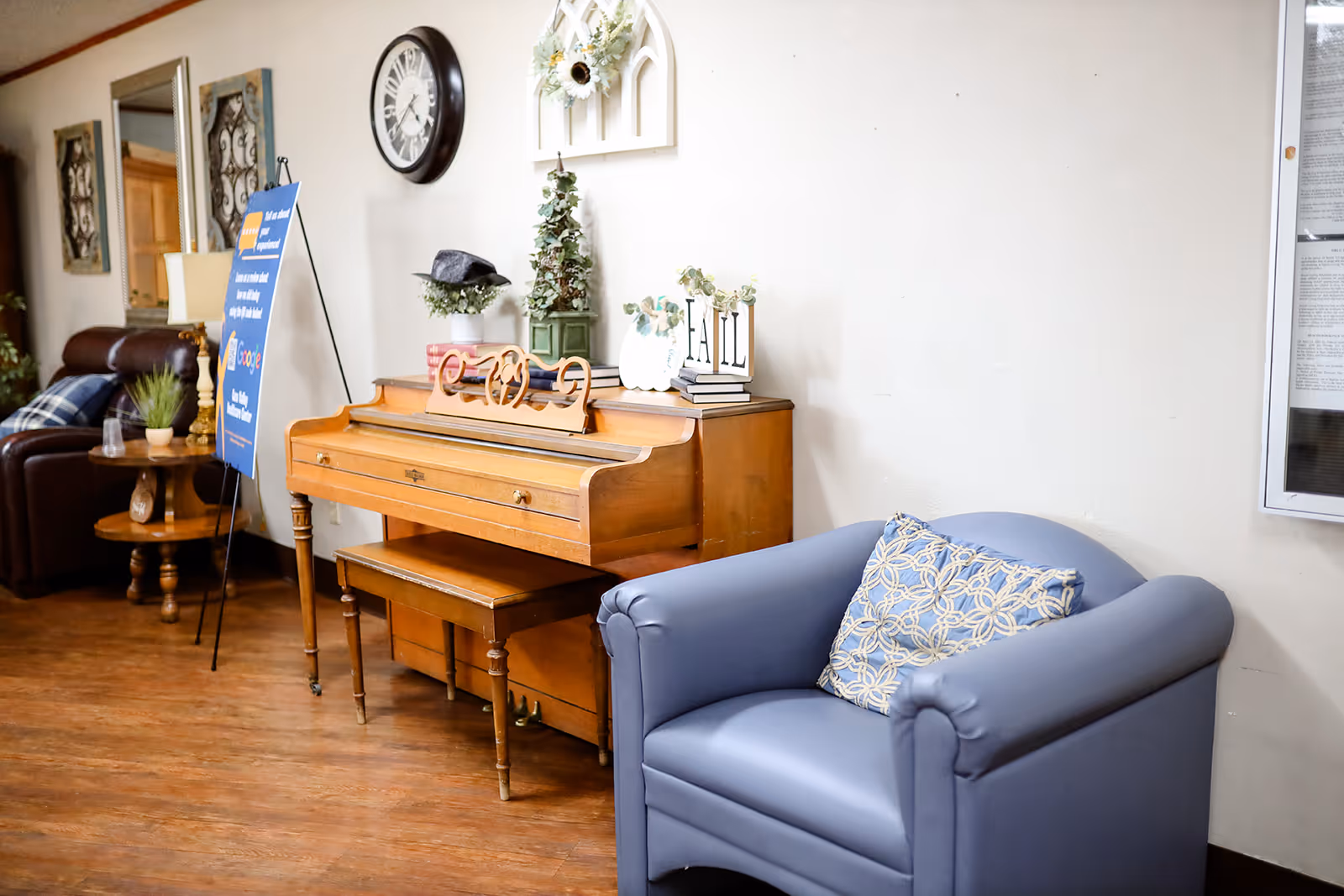 A cozy interior space featuring a wooden piano with decorative items on top, a blue armchair with a patterned cushion, a round wooden side table with a plant, a wall clock, and framed wall decorations. The floor is wooden and the walls are light-colored.