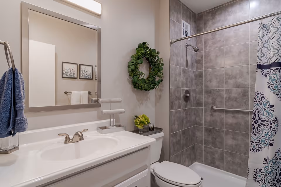 A modern bathroom featuring a white sink with a silver faucet, a large rectangular mirror above the sink, a toilet with a small green plant on the tank, and a shower area with gray tiled walls and a patterned shower curtain. A green wreath hangs on the wall next to the mirror, and a blue towel is hanging on a towel ring beside the sink.