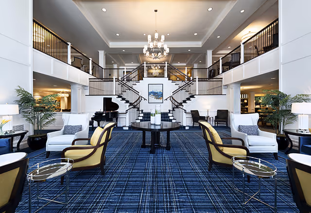 Spacious and elegant senior living facility common area with a blue patterned carpet, multiple seating arrangements including white and yellow armchairs, a grand piano on the left, and a central round table with decorative vases. A large chandelier hangs from the ceiling, and a double staircase with black railings leads to an upper level with additional seating and lighting.