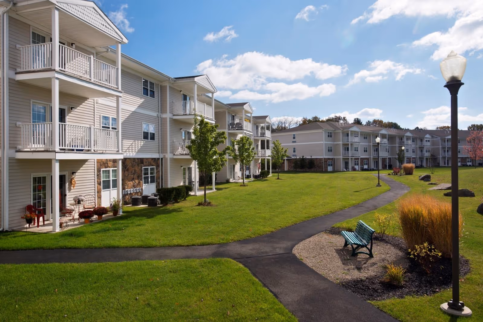 Exterior view of a senior living facility with multiple three-story buildings featuring balconies. A paved walking path winds through a well-maintained grassy area with small trees, benches, and lamp posts under a partly cloudy blue sky.