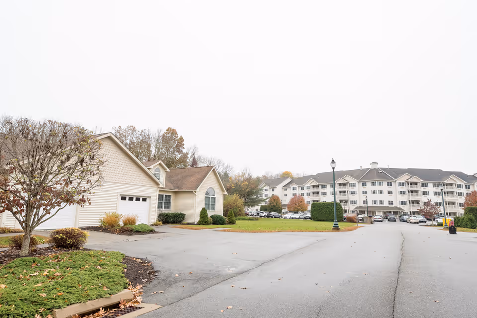 Exterior view of senior living buildings and nearby garages with a driveway, parking area, lamp posts, and landscaping on an overcast day.