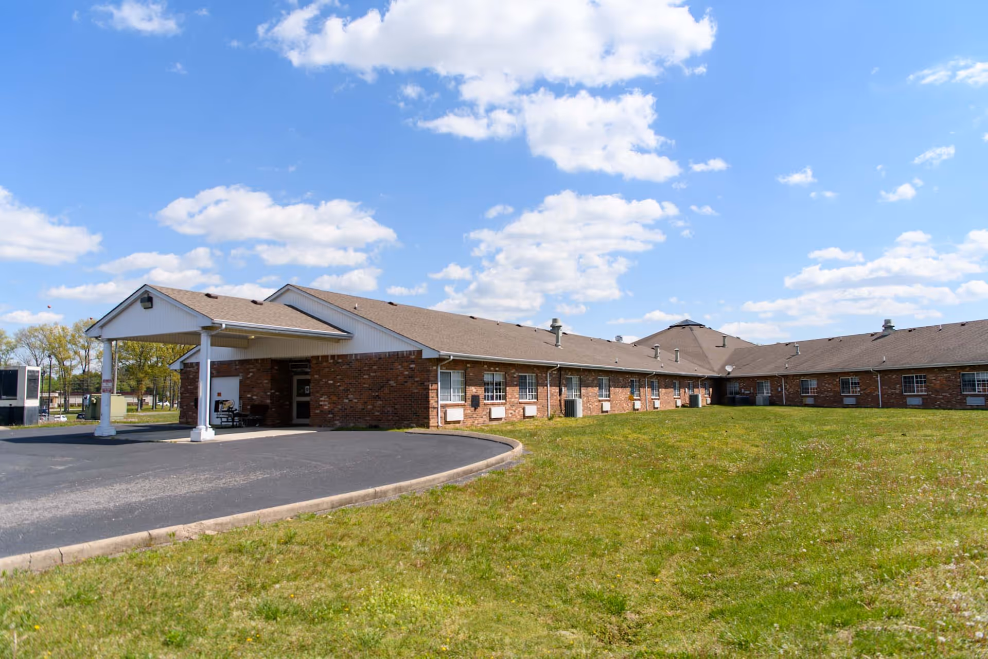 Exterior view of a single-story brick building with a covered entrance and a paved driveway. The building is surrounded by a grassy area under a partly cloudy blue sky.