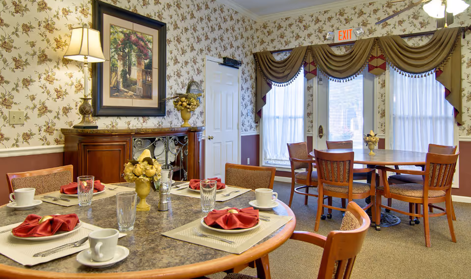 A dining room in a senior living facility with two round tables set for meals. Each table has chairs around it, place settings with white cups, glasses, silverware, and red folded napkins. The room features floral wallpaper, a framed painting, a lamp on a wooden cabinet, and large windows with sheer curtains and decorative valances. An exit sign is visible above a door in the background.