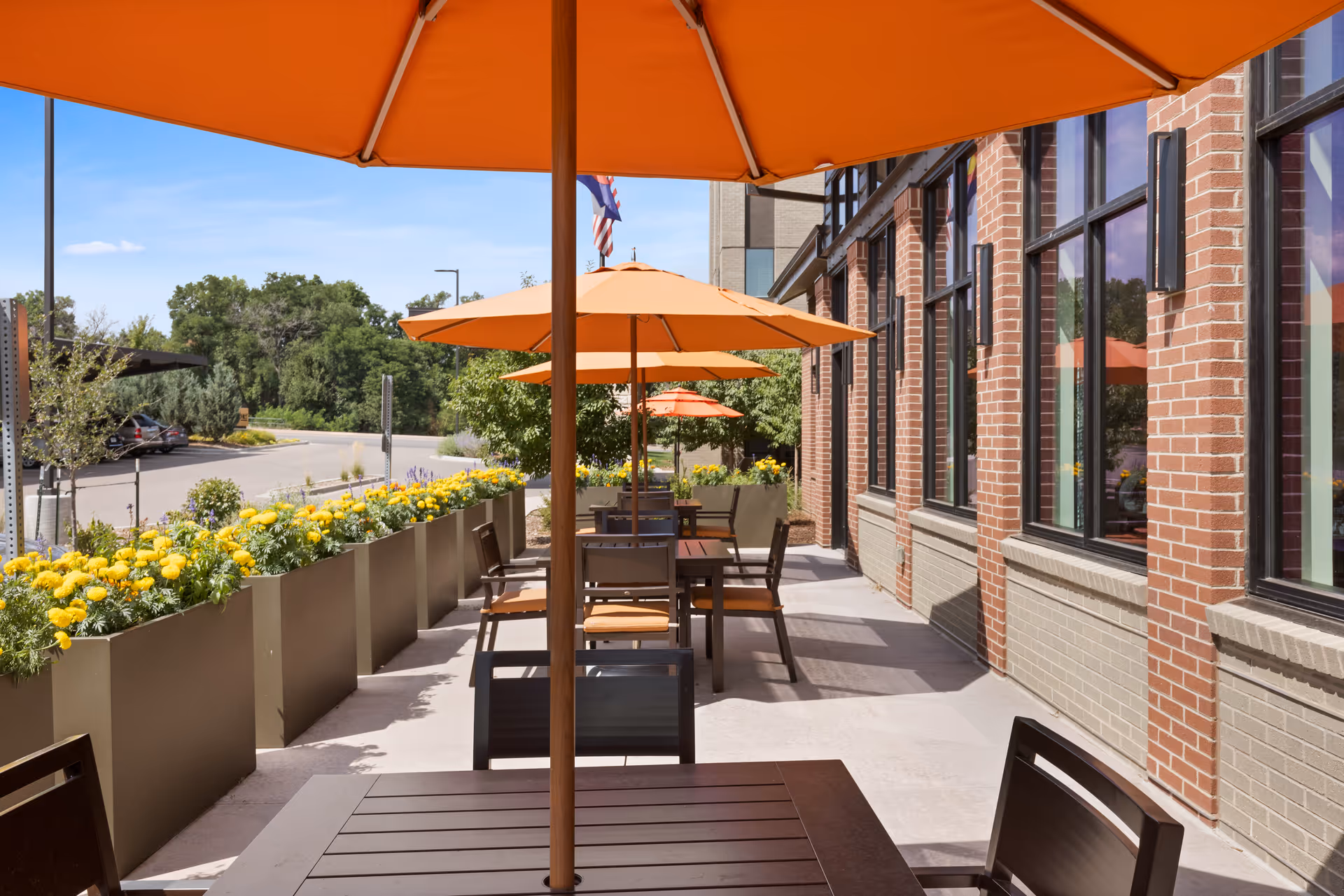 Outdoor patio area at Hyland Hills Senior Living with several tables and chairs under large orange umbrellas. The patio is lined with planters filled with yellow flowers and is adjacent to a brick building with large windows. Trees and a parking area are visible in the background under a clear blue sky.