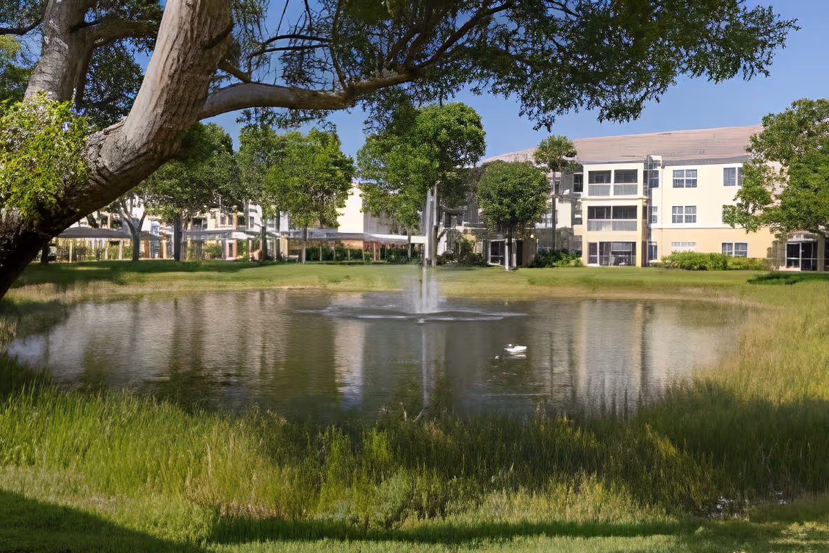 A serene outdoor scene at Aston Gardens At Pelican Marsh featuring a small pond with a central water fountain, surrounded by green grass and trees. In the background, there is a multi-story building with large windows under a clear blue sky.