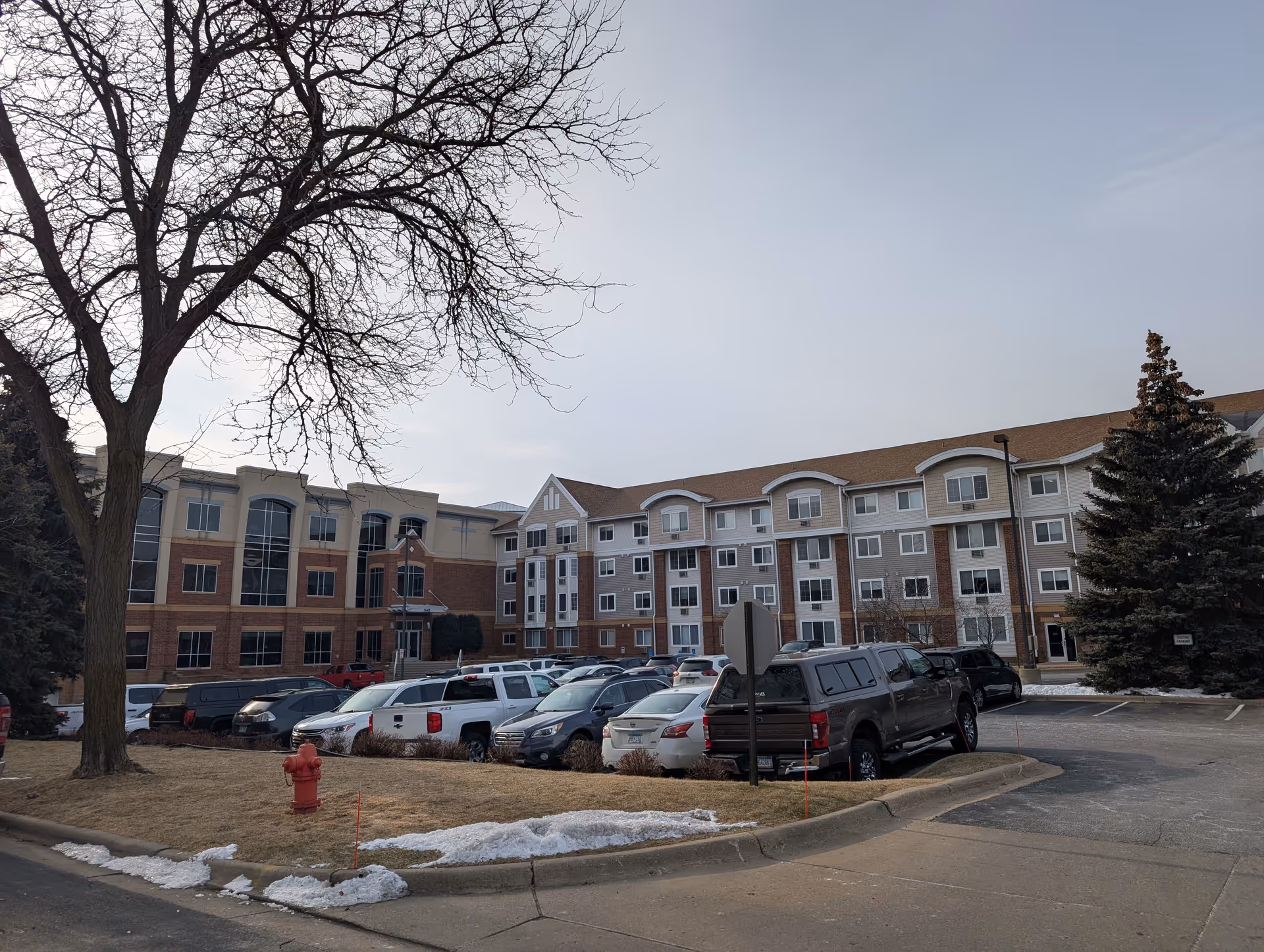 Exterior view of a multi-story senior living facility named Mainstreet Village with a parking lot in front, several parked cars, leafless trees, patches of snow on the ground, and a clear sky.
