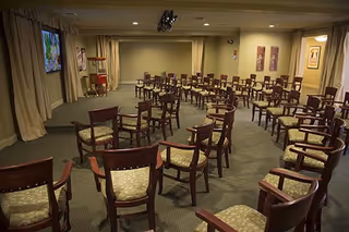 A large room set up with multiple rows of wooden chairs with cushioned seats arranged facing a screen on the left wall. The room has beige walls, carpeted floor, and curtains covering windows. There are framed pictures on the walls and ceiling lights illuminating the space.