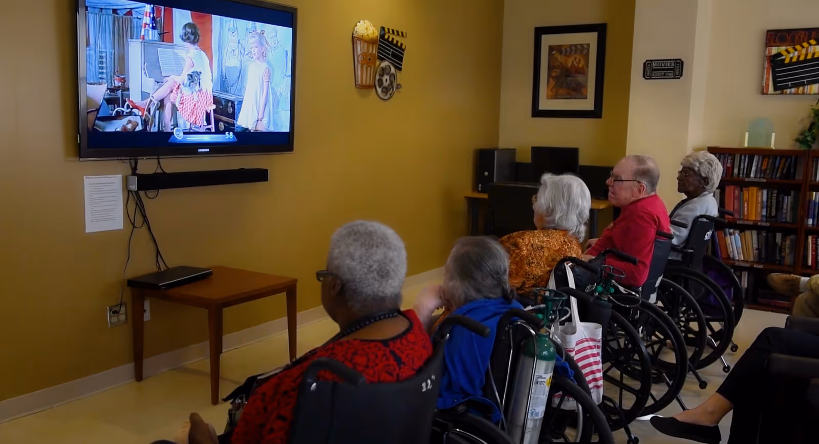 Several elderly residents in wheelchairs seated in a common room watching a movie on a wall-mounted TV.