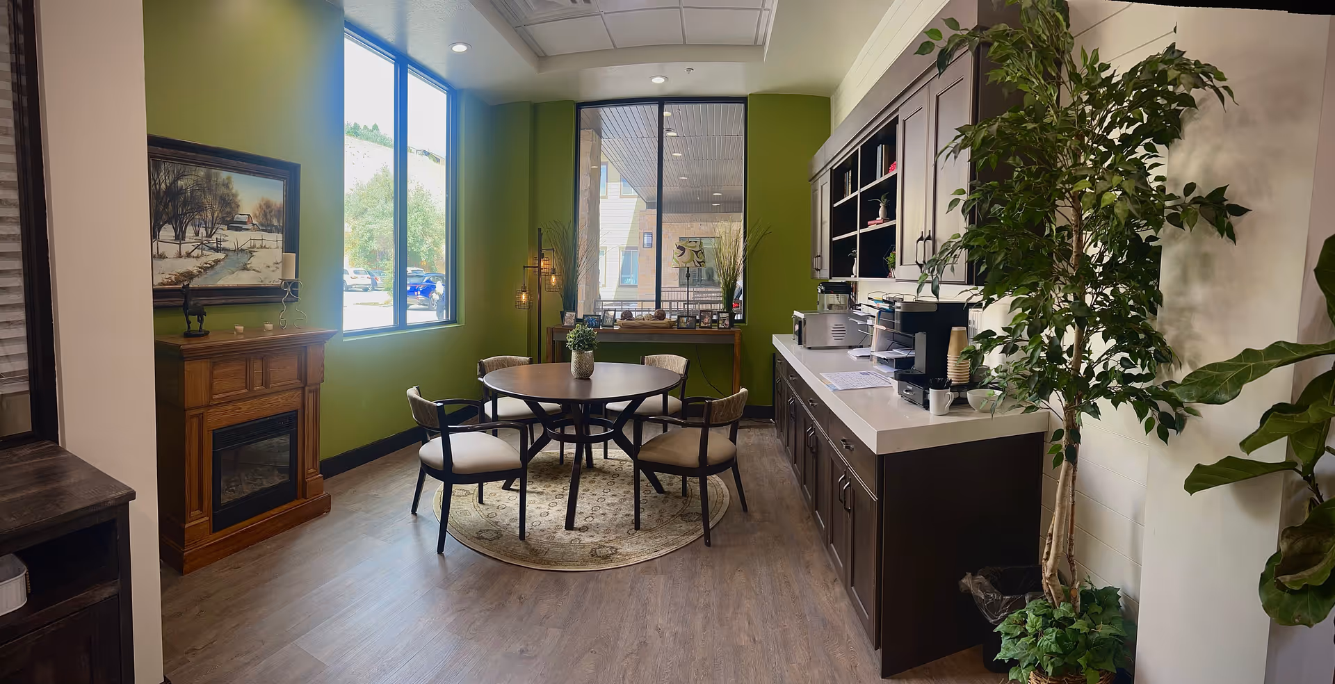 A cozy interior room with green walls featuring a round table with four chairs on a circular rug, a wooden electric fireplace with a painting above it, large windows letting in natural light, a countertop with a coffee machine and cups, and several potted plants adding greenery to the space.