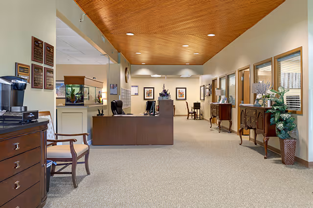 Interior view of a senior living facility reception area with a wooden ceiling, beige walls, and carpeted floor. There is a dark wooden reception desk with a chair behind it, a fish tank on a stand, framed artwork on the walls, and several wooden furniture pieces including chairs and cabinets with decorative plants and flowers.