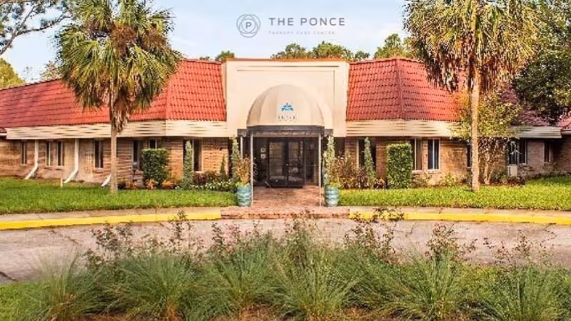 Exterior view of The Ponce Therapy Care Center building with a red roof, beige walls, and a covered entrance. There are palm trees and green shrubs around the building, and a circular driveway in front.