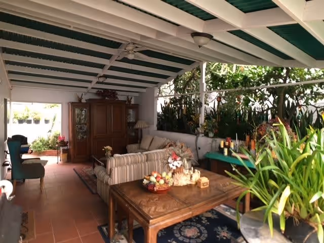 Covered outdoor patio area with a striped sofa, wooden coffee table with decorative items, wooden cabinet, various plants, and a green and white striped ceiling.