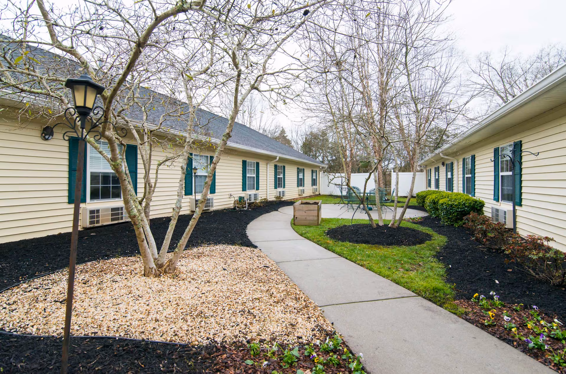 Outdoor courtyard area at Morning Pointe of Clinton with a curved concrete walkway, small trees, landscaped flower beds with mulch and rocks, and patio furniture including a table and chairs. The courtyard is surrounded by single-story buildings with beige siding and green shutters on the windows.