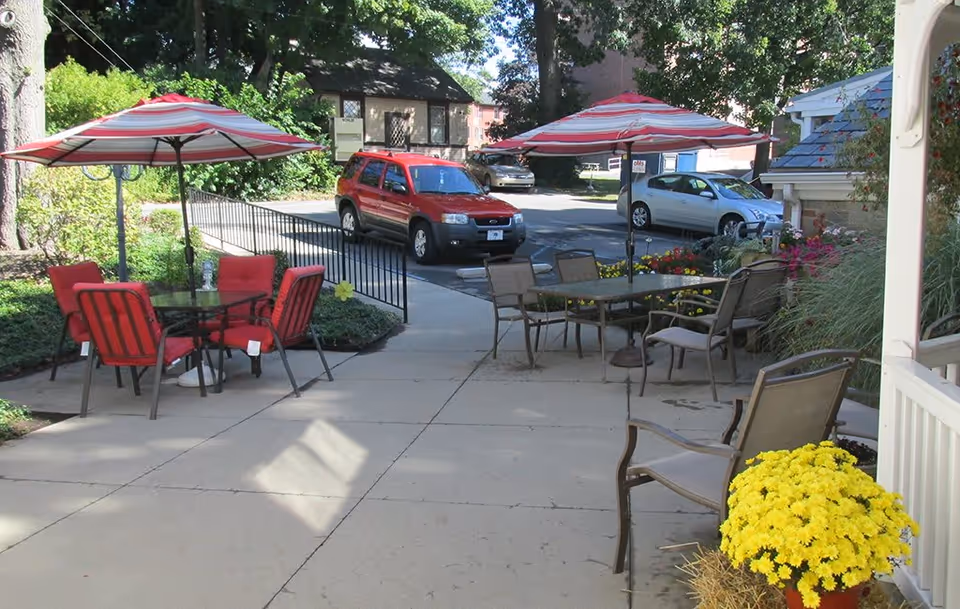 Outdoor patio area with two tables, each shaded by red and white striped umbrellas. One table has four red cushioned chairs, and the other has four beige chairs. There are yellow flowers in a pot in the foreground and some parked cars and greenery in the background.