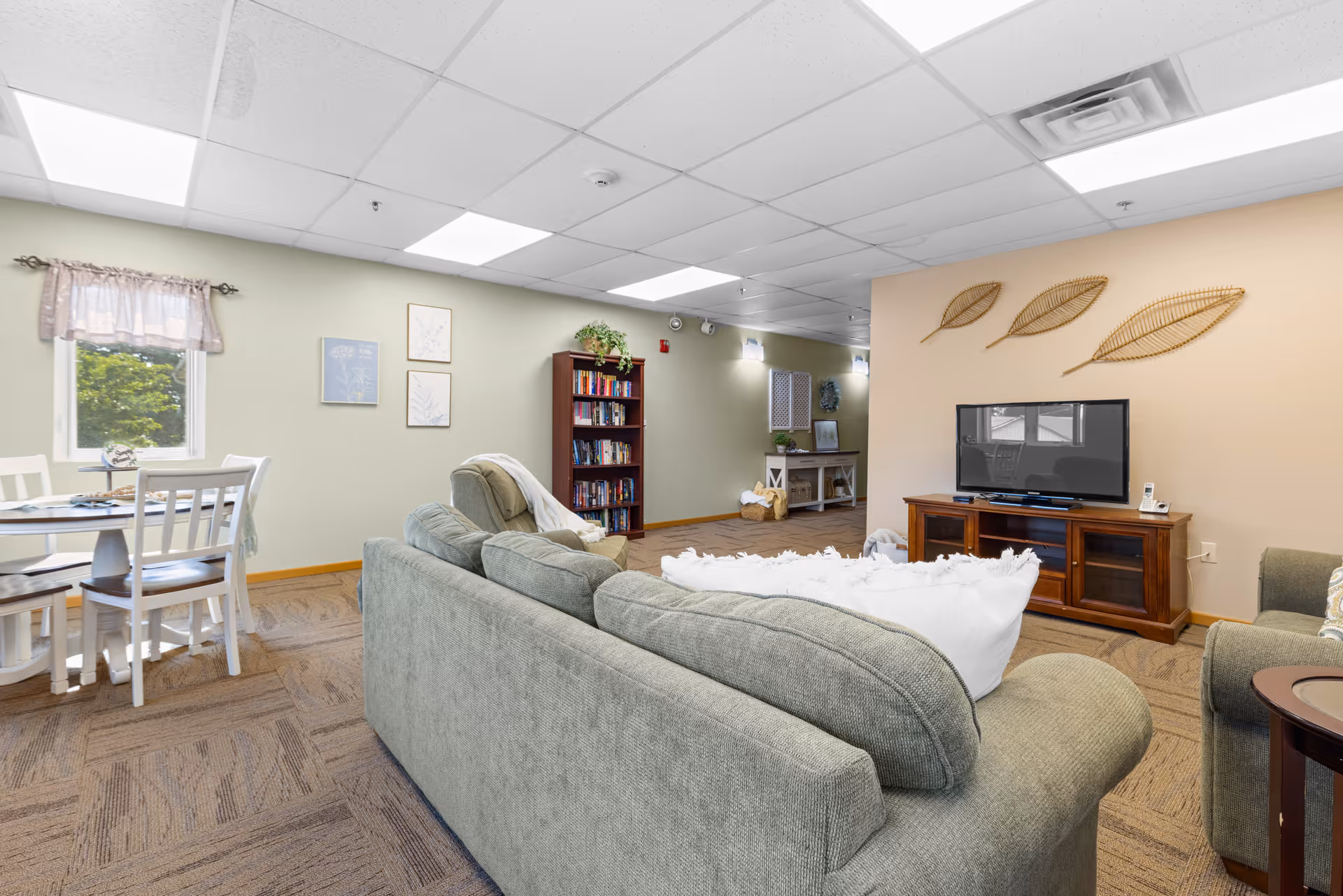 A cozy living room area in Prairie Crossing Assisted Living featuring a green sofa with white pillows, a wooden TV stand with a flat-screen television, and three decorative leaf wall hangings above the TV. To the left, there is a small dining area with a round white table and four chairs near a window with a sheer curtain. A bookshelf filled with books and a small plant on top is against the wall, and the room has beige carpeting and a drop ceiling with fluorescent lighting.